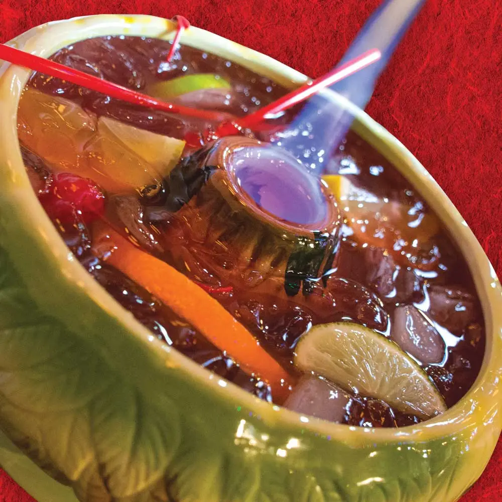 A flaming Volcano Bowl cocktail in a ceramic bowl w/citrus, cherries and long straws